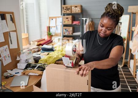 Moderna lavoratrice di magazzino donna che sigilla scatola di cartone con cellotape per posto di lavoro prima di inviare l'ordine imballato al cliente Foto Stock