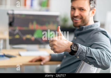 Un uomo d'affari maturo e positivo che lavora al computer con grafici e grafici sullo schermo, mostrando il pollice in su, fuoco selettivo Foto Stock