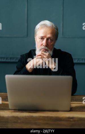 Ritratto verticale del fidato uomo d'affari maturo seduto a tavola di legno con computer portatile in ufficio a casa guardando la fotocamera. Foto Stock