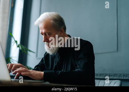 Vista laterale ravvicinata di un uomo d'affari anziano concentrato che lavora su un laptop sul posto di lavoro in una stanza d'ufficio buia. Foto Stock