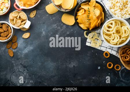 Cibo malsano. Spuntini. Tutti i classici spuntini di patate con arachidi, anelli di popcorn e cipolle e pretzel salati in ciotole su backgroun di cemento nero Foto Stock