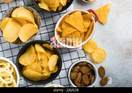 Spuntini. Cibo malsano. Tutti i classici spuntini di patate con arachidi, anelli di popcorn e cipolle e pretzel salati in ciotole su fondo grigio di cemento Foto Stock