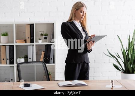 biondo avvocato scrittura sulla clipboard mentre in piedi in ufficio Foto Stock