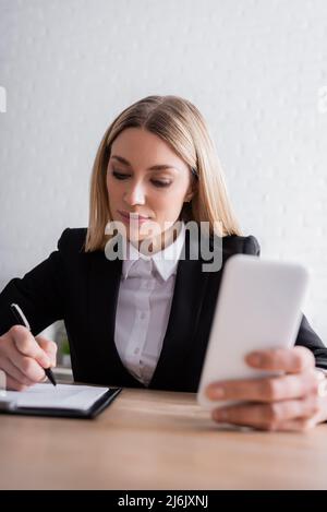 avvocato che tiene lo smartphone e la scrittura in notebook mentre lavora in ufficio Foto Stock