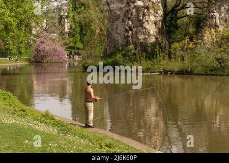 Parigi, Francia - 04 10 2022: Park des Buttes Chaumont. Un pescatore lungo il lago con alberi colorati ed edifici dietro Foto Stock
