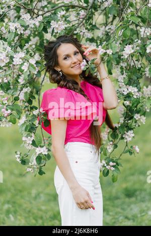 Una giovane donna carina con un pigtail sorride e tiene un ramo fiorente di un albero di mele nel giardino primaverile Foto Stock