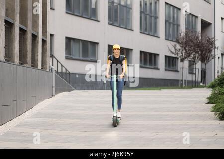 Lady Delivery lavoratore con zaino giallo e berretto corre scooter elettrico lungo la strada della città contro gli edifici. La consegna del cibo del corriere corre la strada su uno scooter elettrico, ordine di consegna in linea Foto Stock