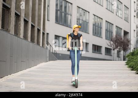 Lady Delivery lavoratore con zaino giallo e berretto corre scooter elettrico lungo la strada della città contro gli edifici. La consegna del cibo del corriere corre la strada su uno scooter elettrico, ordine di consegna in linea Foto Stock
