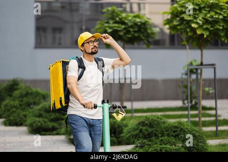 Consegna ragazzo in berretto giallo su scooter elettrico in viaggio, consegna on-line di cibo e generi alimentari ordini ai clienti. Corriere di cibo, servizio di consegna. Foto Stock