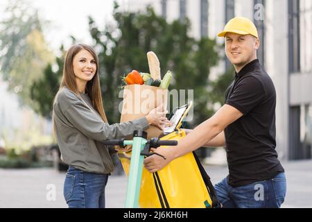 Cliente donna caucasica che prende la consegna carta eco sacchetto con la spesa takeout cibo pasto da uomo corriere che tiene carta pacchetto consegna supermercato o ristorante takeaway ordine in piedi all'aperto Foto Stock