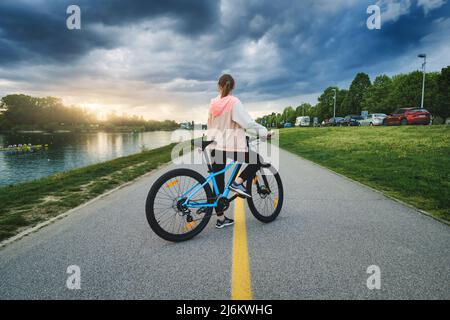 Donna che guida una mountain bike sulla strada vicino al lago al tramonto Foto Stock