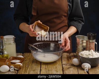Cucinare l'impasto in una ciotola di vetro per mano di uno chef professionista su un tavolo da cucina in legno. Uova, farina e altri ingredienti sul tavolo. Primo piano. Foto Stock