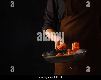 Uno chef professionista prepara pezzi di pesce rosso - tonno, salmone, trota in una padella su sfondo nero. Levitazione. Il concetto è ricette per il pesce e. Foto Stock