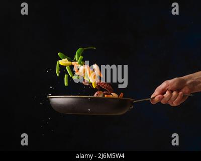 Uno chef professionista prepara frutti di mare - gamberi e polpi con pezzi di verdure in una padella su sfondo scuro. Cibo congelato a bordo. Healt Foto Stock