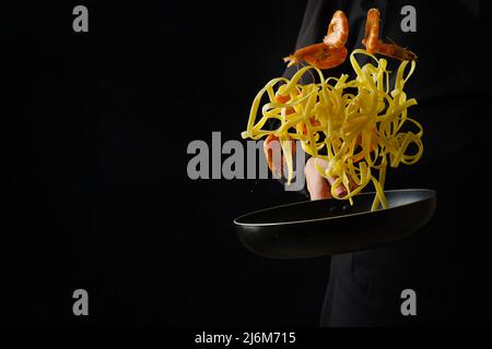 Cucina italiana. Gamberetti la pasta in una padella su sfondo nero in un volo congelato in mano di uno chef professionista in uniforme nera. Pubblicità Foto Stock