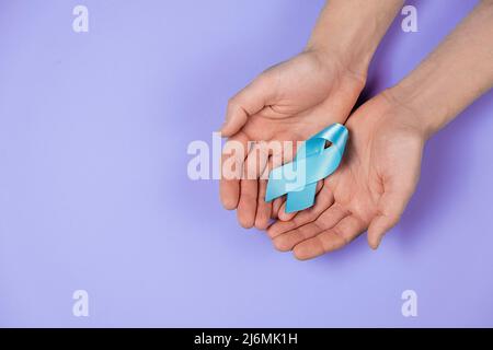 Primo piano vista dall'alto delle mani maschili che tengono un piccolo nastro di consapevolezza blu sulla parete di fondo di colore viola in studio con spazio per la copia per la pubblicità. Symbo Foto Stock