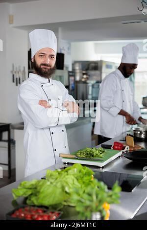 Positivo sous chef taglio erbe fresche da utilizzare in piatti della cucina gourmet mentre si guarda la macchina fotografica. Esperto di gastronomia che indossa uniforme da cucina mentre prepara guarnire per il servizio cena. Foto Stock