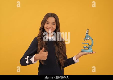 happy kid tenere microscopio per l'istruzione scolastica su sfondo giallo mostrando pollice su, biologia Foto Stock