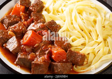 Carne di manzo speziata lentamente in vino rosso con verdure servite con tagliatelle in primo piano in un piatto sul tavolo. Orizzontale Foto Stock