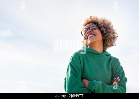 giovane donna latina con occhiali e capelli afro sorridendo felice con il cielo sullo sfondo, concetto di felicità e stile di vita, copia spazio per il testo Foto Stock