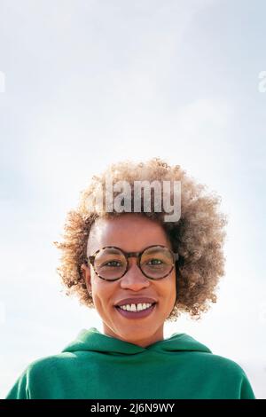 ritratto verticale di una giovane donna latina con occhiali e capelli afro sorridenti felici e guardando la macchina fotografica con il cielo sullo sfondo, concetto di felicità a. Foto Stock