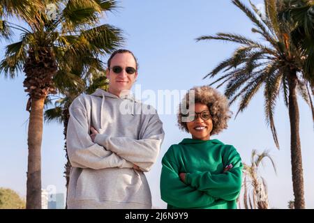 coppia di amici con le braccia incrociate sorridendo felice guardando la macchina fotografica accanto alle palme, concetto di amicizia e rapporto di coppia Foto Stock