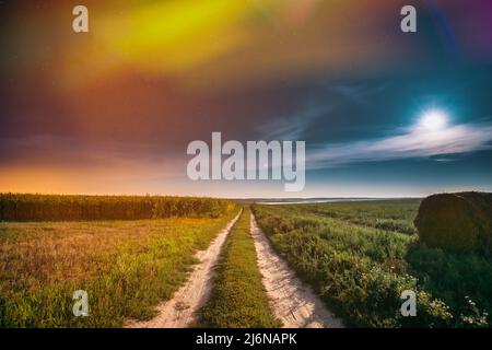 Notte Starry Sky sopra Country Road attraverso Field Meadow con Hay Bale dopo Harvest. Stelle incandescenti e Luna piena sopra il paesaggio rurale. Colorato Foto Stock