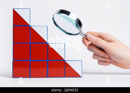 Mano con lente d'ingrandimento sopra la carta cadente dai cubetti. Analisi dei motivi e delle conseguenze del fallimento e della crisi economica. Ricerca e pianificazione di modi per risolvere i problemi. Foto Foto Stock