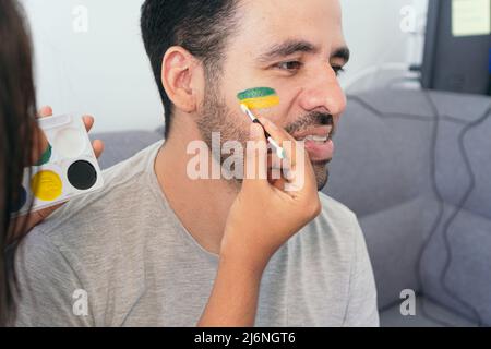 Donna che dipinge la bandiera brasiliana sul volto di un amico Foto Stock