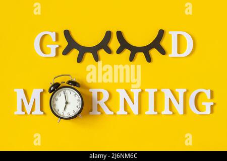 Concetto creativo di buon mattino. Lettere bianche, palpebre nere e sveglia su sfondo giallo. Vista dall'alto, disposizione piatta. Biglietto di auguri. Foto Stock
