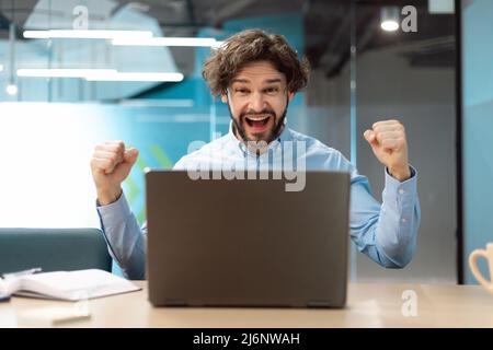 Un uomo d'affari entusiasta che utilizza il pc festeggia il successo scuotendo i pugni urlando sì Foto Stock