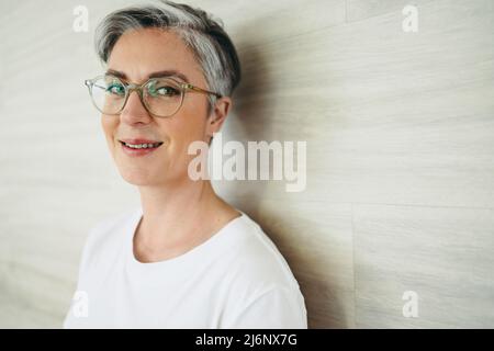 Ritratto di una donna d'affari matura sorridendo alla macchina fotografica mentre indossa gli occhiali. Donna d'affari felice in piedi contro un muro in un ambiente di lavoro moderno. Foto Stock