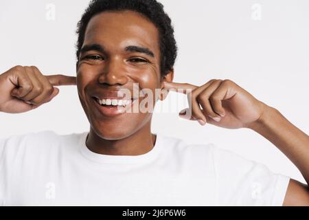 Giovane uomo nero in t-shirt sorridente mentre gli si tappano le orecchie isolato su sfondo bianco Foto Stock