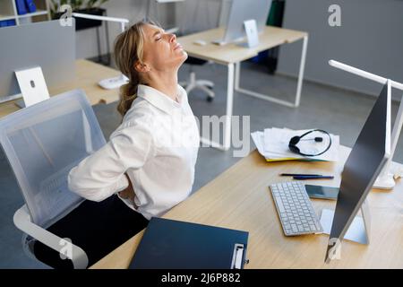 Donna d'affari di mezza età che ha dolore alla schiena mentre si siede alla scrivania d'ufficio Foto Stock