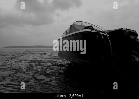 Una foto monocromatica di una barca rivolta verso il mare, bloccata nel fango su una spiaggia Foto Stock