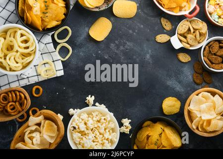 Cibo malsano. Spuntini. Tutti i classici spuntini di patate con arachidi, anelli di popcorn e cipolle e pretzel salati in ciotole su backgroun di cemento nero Foto Stock