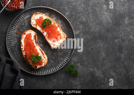 Due dandwiches con caviale rosso. Caviale rosso salmone in ciotola e sandwich server su vecchio piatto di ferro su sfondo vecchio tavolo nero. Vista dall'alto. Spazio di copia. Foto Stock