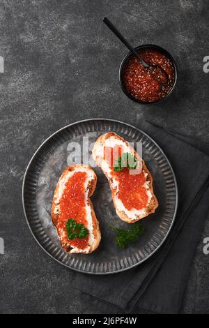 Due dandwiches con caviale rosso. Caviale rosso salmone in ciotola e sandwich server su vecchio piatto di ferro su sfondo vecchio tavolo nero. Vista dall'alto. Spazio di copia. Foto Stock