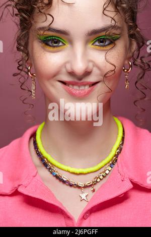 Ritratto di giovane donna sorridente vestita in abito rosa su sfondo rosa. Donna con insoliti occhi verdi ombre trucco, capelli ricci e orecchini. Foto Stock