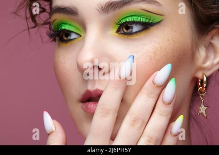 Ritratto di giovane donna con mano vicino al viso. Femmina con insoliti occhi verdi ombre trucco. Ragazza con chiodi con manicure multicolore. Foto Stock