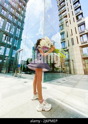 Modella donna in giovane fit con bouquet di delphinium bianco e viola in piedi in abito corto e scintillante punteggiato. Altezza completa Foto Stock