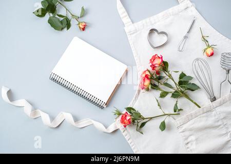 Libro da cucina in bianco, accessori da forno con grembiule con fiori rose su sfondo grigio, piatto, vista dall'alto. Ricette festive di cottura concetto. Foto Stock