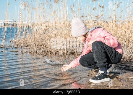 Ragazza affascinante in abiti caldi gioca sulla riva del lago. Giorno fresco e soleggiato. Cielo blu e sfondo di canne secche. Attività all'aperto Foto Stock