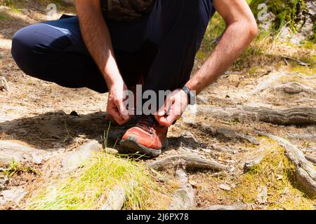 camminatore maschio adulto accovacciato in pantaloni blu e un primo piano delle sue mani che legano i lacci delle sue sneakers rosse su alcune radici in una foresta, dopo aver camminato alo Foto Stock