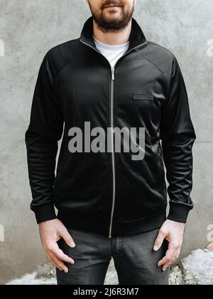 Giovane uomo con una barba in una giacca sportiva nera. Vista frontale su sfondo di pareti in cemento grigio chiaro. Design e layout di giacca uomo sportiva uomo da uomo per stampa. Foto Stock