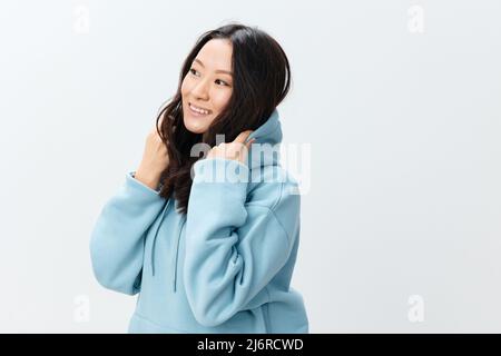 Alla moda sorridente felice ragazza asiatica giovane donna in felpa blu mette sul cappuccio in posa isolata su sfondo bianco studio. La migliore offerta Foto Stock