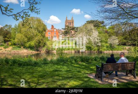 Due adulti seduti su una panchina del parco che si affaccia sulla Cattedrale di Hereford dalle rive del fiume Wye, Hereford, Inghilterra, Regno Unito Foto Stock