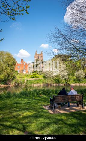 Due adulti seduti su una panchina del parco che si affaccia sulla Cattedrale di Hereford dalle rive del fiume Wye, Hereford, Inghilterra, Regno Unito Foto Stock