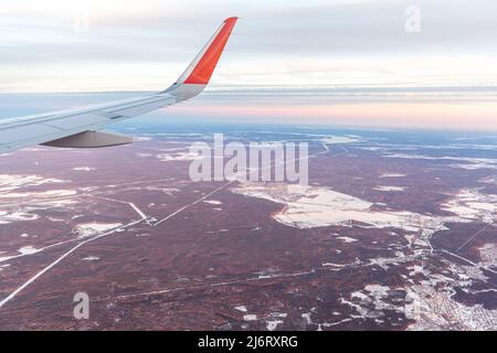 Volo in aereo al tramonto o all'alba. L'ala e la terra dell'aeromobile sono visibili attraverso l'illuminatore. Vista dalla finestra del piano. Aereo, aereo. Traversata Foto Stock