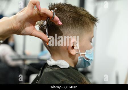 Un ragazzo adolescente ottiene un taglio di capelli in un barbiere durante una pandemia, un taglio di capelli nel salone, un cliente e un parrucchiere in maschere, un taglio di capelli del bambino con sciss Foto Stock